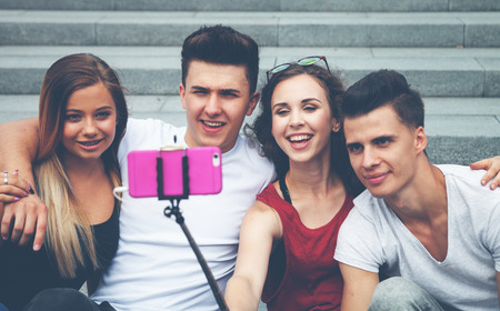 Group of friends sitting together with mobile phone on selfie stick and taking pictureの写真素材