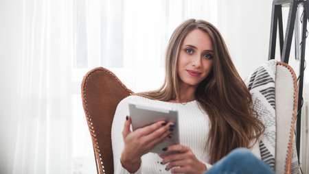 Young woman at home sitting on modern chair and using tablet computer, relaxing in living room