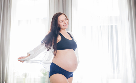 Happy and proud smiling pregnant woman at home dancing near windowの写真素材