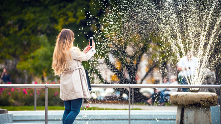 Woman in the autumn city taking picture of fountain using smartphoneの写真素材