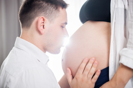 Young father kissing pregnant belly of his wife at homeの写真素材