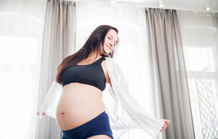 Happy and proud smiling pregnant woman at home dancing near windowの写真素材
