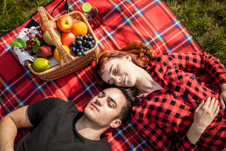 Loving young couple lying on picnic blanket near basket full of fruits enjoying sunny dayの写真素材