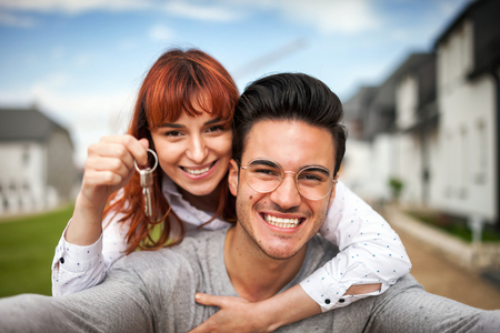 Smiling couple with keys to their new home hugging and looking at camera taking selfieの写真素材