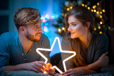 Couple in love at Christmas evening holding glowing white LED star, cozy dark living roomの写真素材