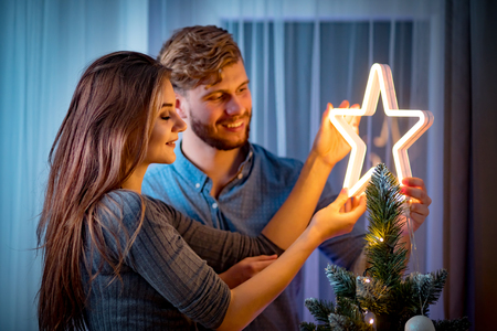 Cheerful couple decorating christmas tree by glowing LED star in dark cozy roomの写真素材