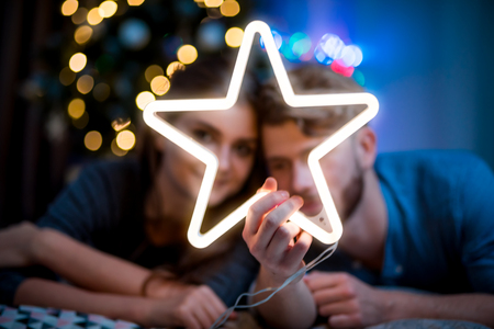Couple in love at Christmas evening holding glowing white LED star, cozy dark living roomの写真素材