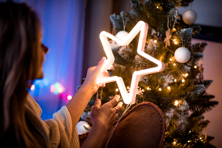 Cheerful young woman sitting next to christmas tree and holding glowing LED star in dark cozy room at xmas eveの写真素材