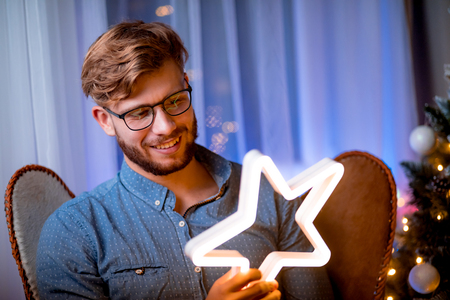 Cheerful handsome man sitting next to christmas tree and holding glowing LED star in cozy room at xmas eveの写真素材