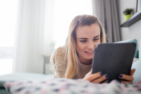 Happy woman reading ebook on digital reader while lying on comfortable sofa at home, leisure time