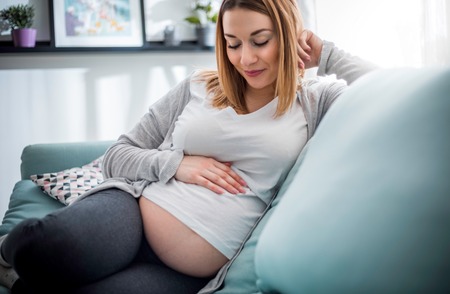 Pregnant woman relaxing on sofa at home in sunny living roomの写真素材