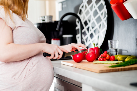 Pregnant woman preparing salad and cutting vegetables in kitchenの写真素材