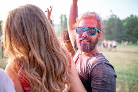 Bearded young man covered in colorful powder standing with friends at holi festival