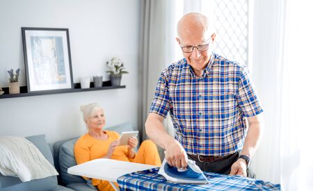 Senior man doing housework with iron and clothes while his wife using tabletの写真素材