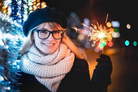 Happy smiling girl holding sparklers standing on illuminated city street during Christmas or New...