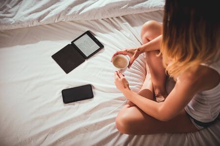 Young woman sitting on bed with cup of coffe using ebook reader tablet and smartphone, top view