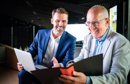 Younger and older businessman talking about documents during meeting in modern business loungeの写真素材