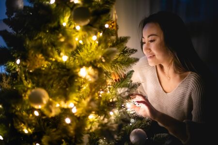Beautiful Asian woman decorating Christmas tree in home at eveningの写真素材
