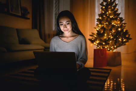Happy asian girl shopping online with laptop sitting near decorated christmas tree background at eveningの写真素材