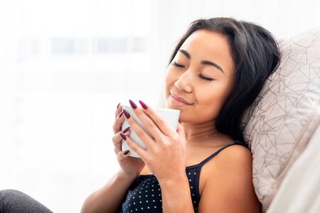 Relaxed young asian woman sitting in armchair with blanket drinking coffee or teaの写真素材
