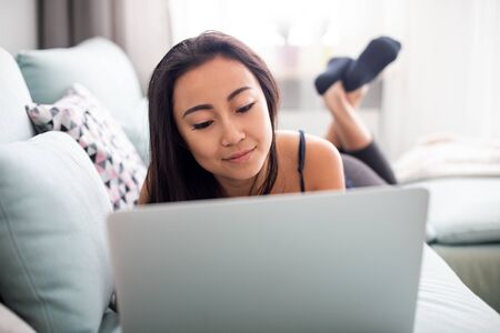 Smiling asian young woman using laptop lying on sofa at homeの写真素材