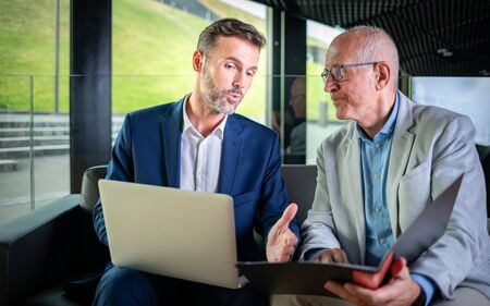 Businessman using laptop to discuss information with older colleague in modern business loungeの写真素材