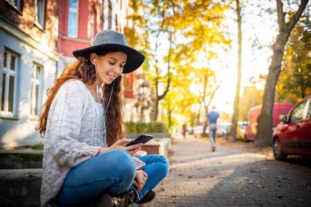 Beautiful stylish woman sitting on street with crossed legs at autumn day and using mobile phoneの写真素材