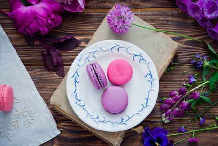 Purple and pink French macaroon cookies on a white plate on a book and a variety of pink and purple flowers on a wooden backgroundの写真素材