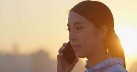 Woman talk to mobile phone in city at sunsetの写真素材