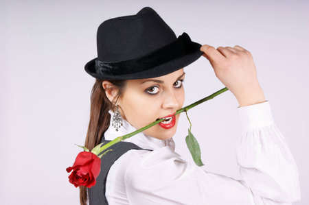 Portrait of a beautiful young woman wearing white blouse, black pinstriped gilet and black hat. The woman is holding a red rose in mouth while looking at camera.の写真素材