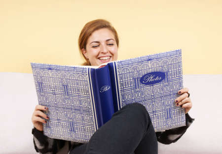 Beautiful young woman sitting on a sofa and looking at photo album. Studio shotの写真素材