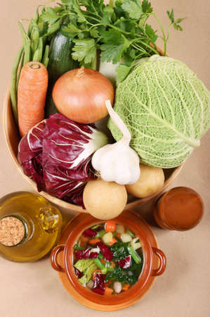Homemade typical italian minestrone (vegetable soup) in a rustic ceramic bowl and vegetables used to prepare it. Studio shot over light brown background.の写真素材