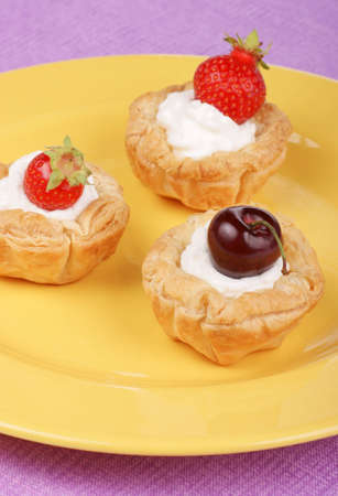 Small tarts with whipped cream, strawberries and cherries served on a yellow plateの写真素材