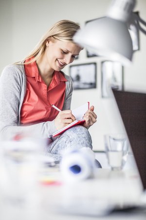 Happy young businesswoman writing in diary at officeの写真素材
