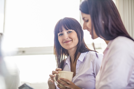 Portrait of young businesswoman holding coffee cup by female colleague in officeの写真素材