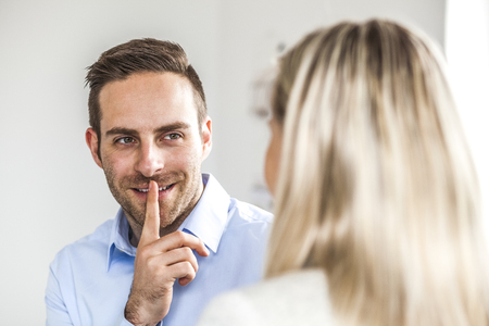 Smiling young businessman gesturing silence while looking at female colleague in officeの写真素材