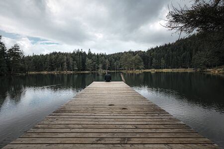 man sitting on the end of a pier jetty looking out to the water in a lake in a forest on a gloomy cloudy dayの写真素材