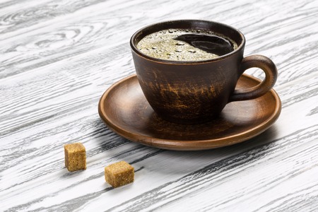Black strong coffee with foam in a ceramic cup on a light wooden table. There are two grains of coffee on a saucer and two cubes of brown sugar.の写真素材