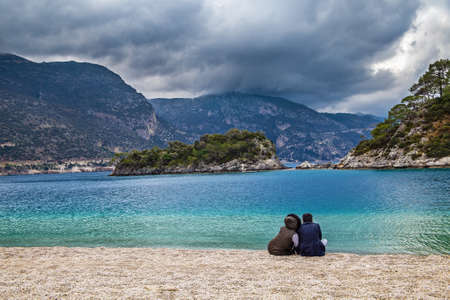 Popular Oludeniz beach on the background of the mountains in pre-thunderstorm weather. There are a couple of people sitting on the pebble shore.の写真素材