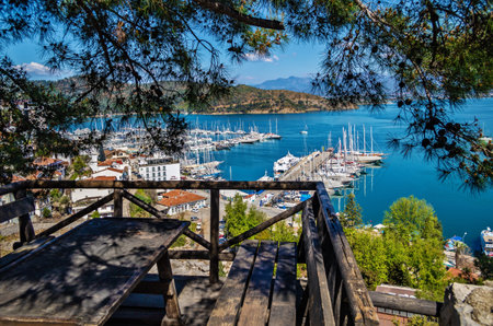 Fethiye, Turkey - March 28, 2021: View of Fethiye harbor with yachts and boats and mountains. There is a wooden table with benches for rest in the foreground. Mugla Province.のeditorial素材