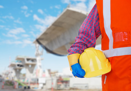 Safety Protective Work Equipment. Worker in orange vest holding yellow helmetagainst construction...