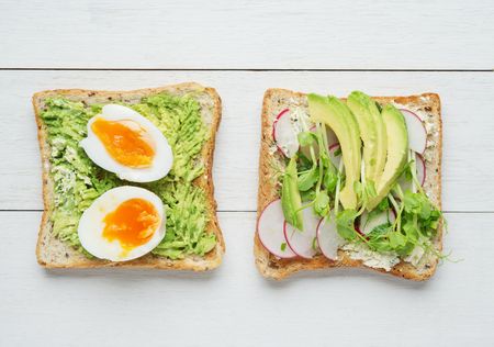 Two avocado toasts with soft boiled eggs,mashed avocado,sliced avocado ,radish, snow pea sprouts and goat cheese on white wooden backgroundの写真素材