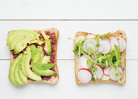 Two avocado toasts with mashed avocado and beetroot,sliced avocado ,radish, snow pea sprouts and goat cheese on white wooden backgroundの写真素材