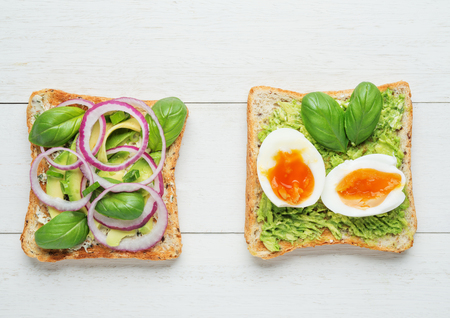 Two avocado toasts with soft boiled eggs,mashed avocado,sliced avocado ,red onion,basil leaves and goat cheese on white wooden backgroundの写真素材