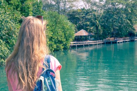 girl with blond long hair stands on the riverbank, hiking with a backpack