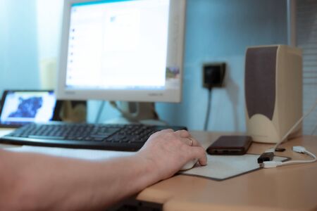 a man sits at home at a table and works on a computer, holds a computer mouse with his hand,...