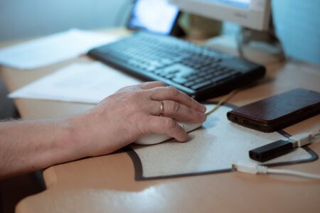 an elderly man sits at a table and works on a computer, holds a computer mouse with his hand, socialization of people of retirement ageの写真素材