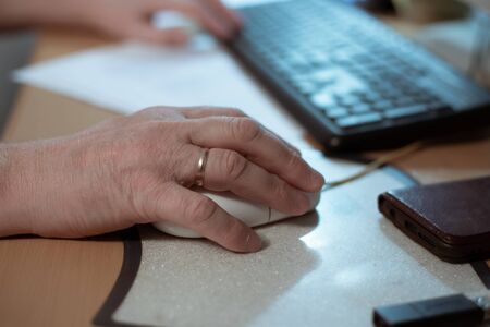 hand of an elderly man lies on a computer mouse, at a computer table, retired at work at homeの写真素材