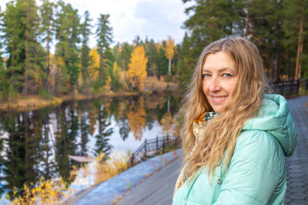 A blonde woman in a jacket stands on the embankment of the river in the autumn forest among tall pines. Spending time in nature.の写真素材