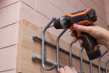 A man with a screwdriver installs hooks for clothes on a brick wall. Repair and home improvement.